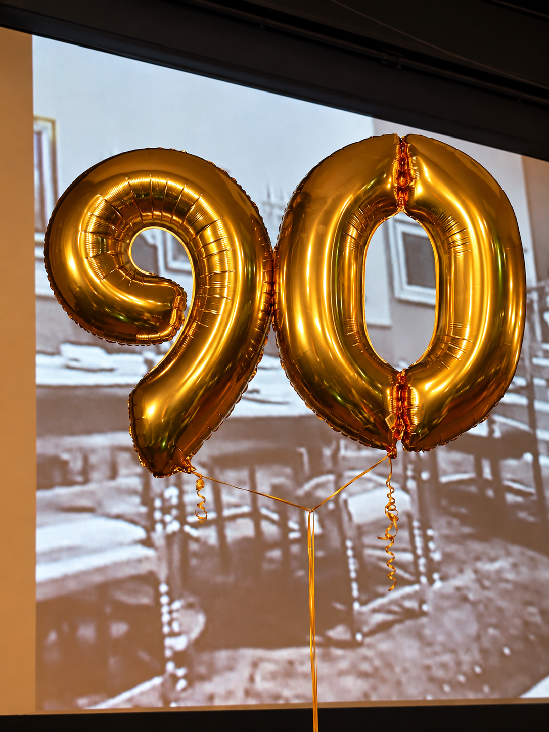 Goldene 90-Jahre-Ballons vor historischen Schwarz-Weiß-Aufnahmen auf der Kinoleinwand.